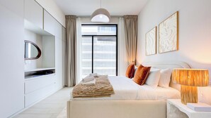 Un dormitorio moderno con una cama grande, una mesita de noche con lámpara, un televisor sobre un mueble bajo y vistas de la ciudad a través de la ventana.