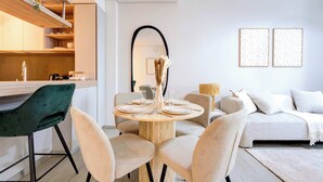 Una sala moderna con una mesa de comedor redonda, sillas color beige, un sofá y una mesa de centro. Se observa un área de cocina con armarios de madera y una planta colgante.