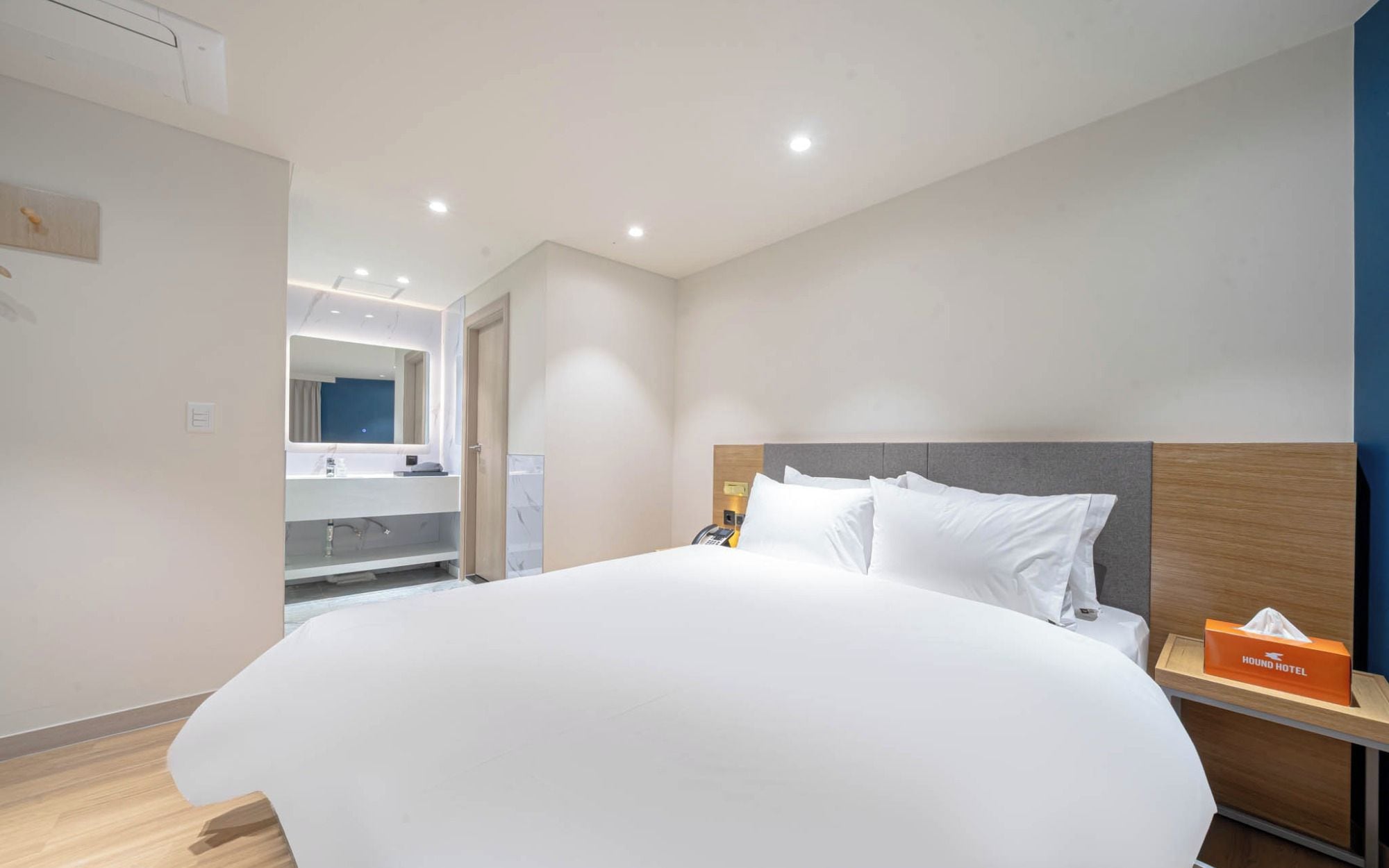 Ein Hotelzimmer mit einem großen Bett, einem Nachttisch mit Taschentuchbox und einem durchgegangenen Türspalte sichtbaren Badezimmer.