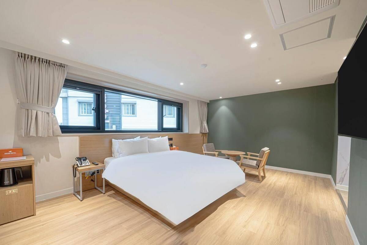 Habitación de hotel moderna con una cama grande, mesita de noche, lámpara y vistas a edificios a través de la ventana.