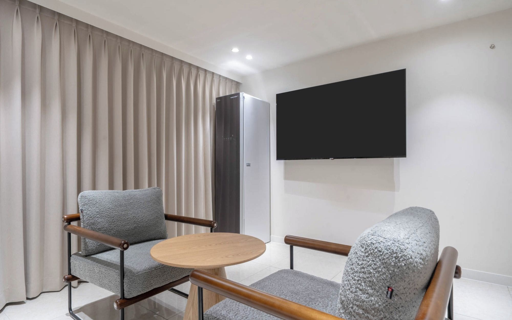 A modern hotel room with a flat-screen TV mounted on the wall, a grey upholstered sofa, a wooden coffee table, and a wardrobe.