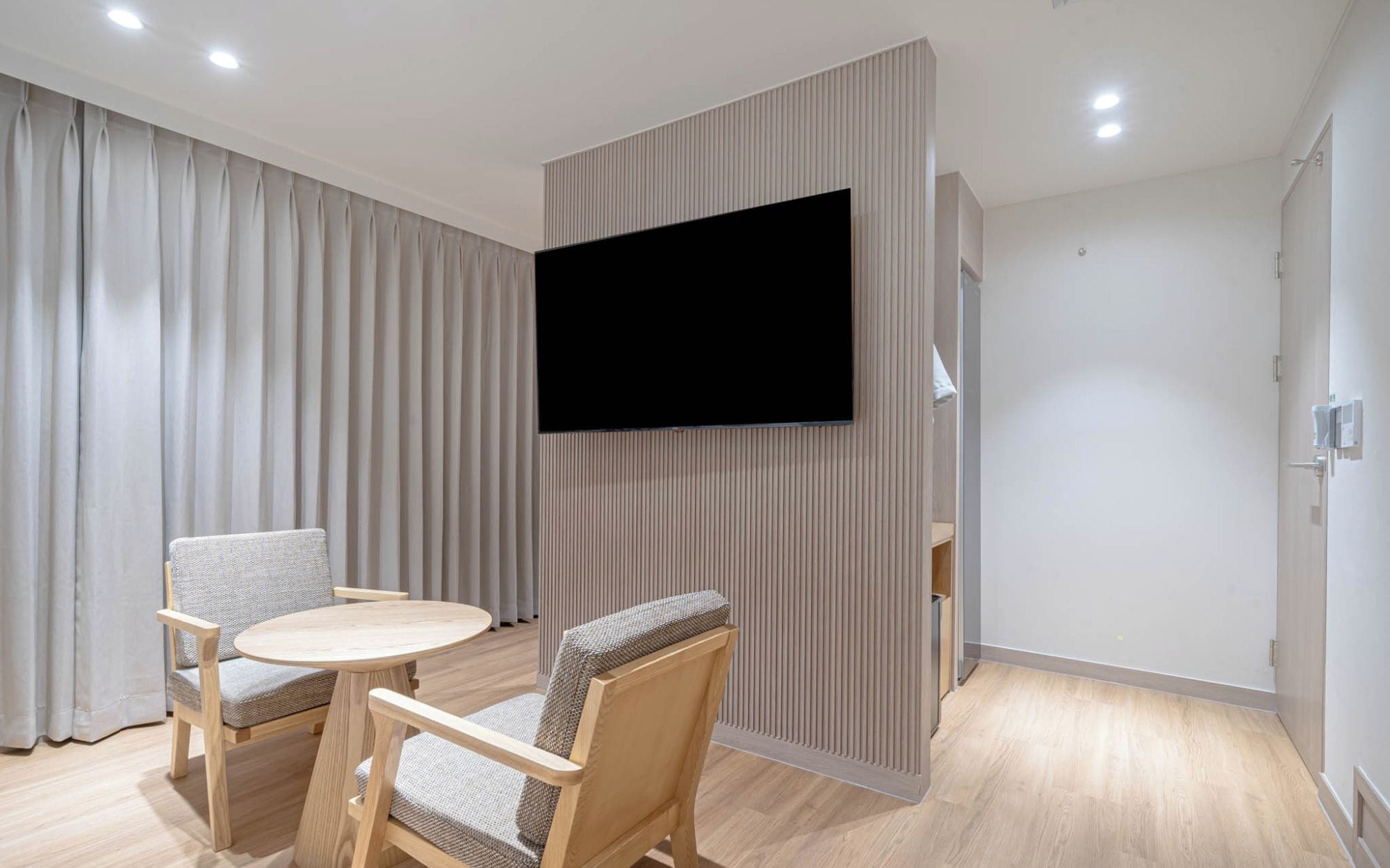Una sala moderna con un televisor de pantalla plana montado en la pared, una mesa redonda de madera, dos sillones tapizados y un ventanal con cortinas.