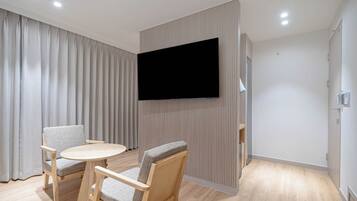 Una sala moderna con un televisor de pantalla plana montado en la pared, una mesa redonda de madera, dos sillones tapizados y un ventanal con cortinas.