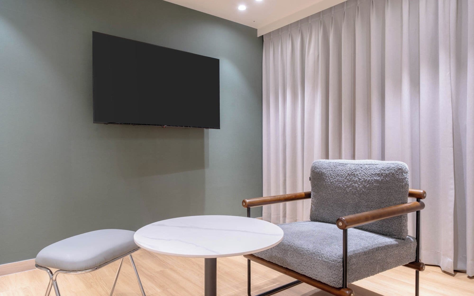 A modern room with a grey armchair, a small round table, a flat-screen TV mounted on the wall, and a window with white curtains.