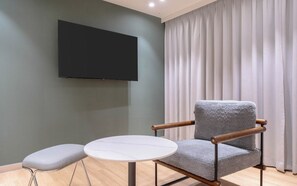 A modern room with a grey armchair, a small round table, a flat-screen TV mounted on the wall, and a window with white curtains. - Ulsan Hound Hotel Samsan (Ulsan)