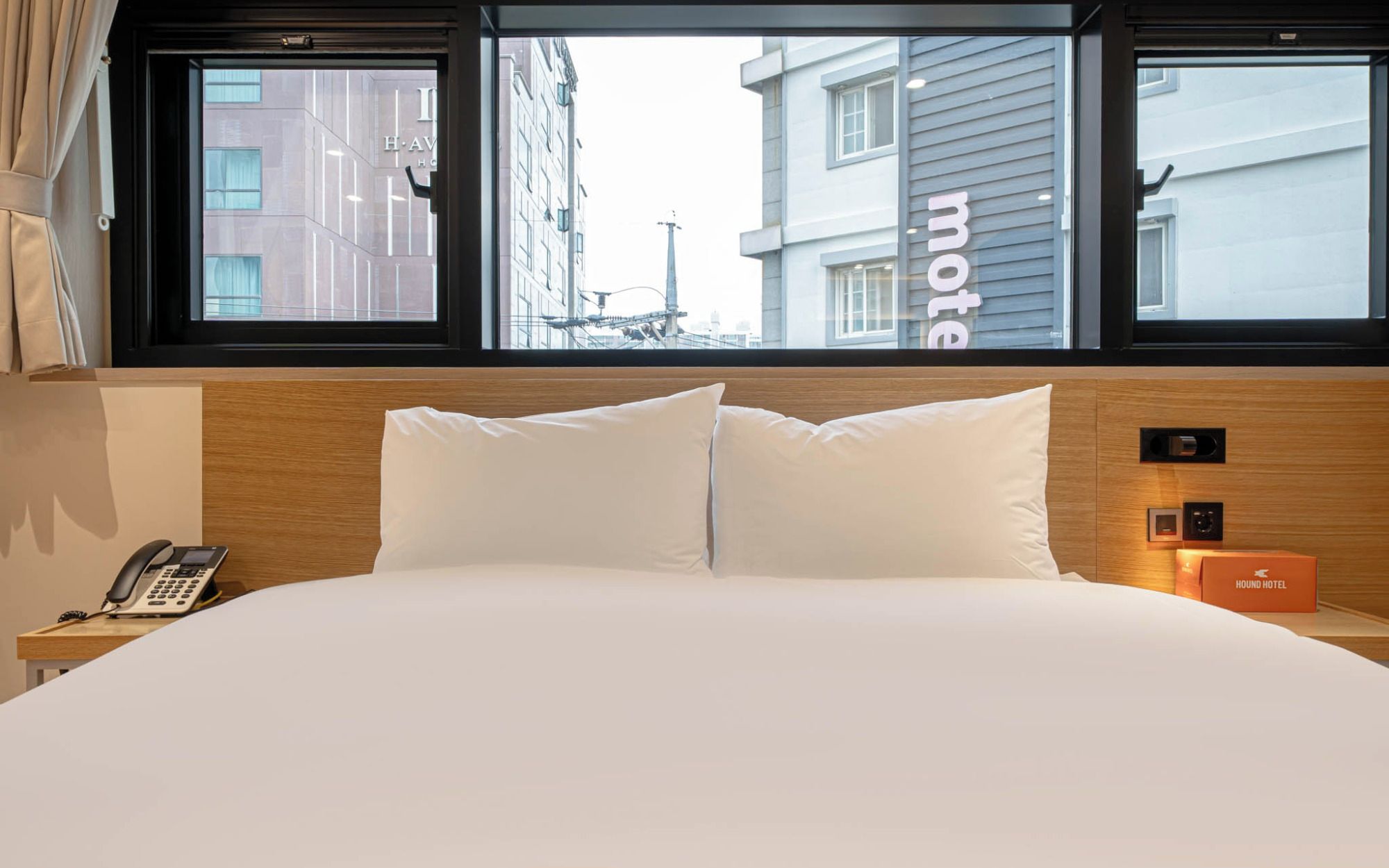 Habitación de hotel con cama, teléfono en la mesita de noche, ventana con vista a edificios y un cartel que indica que es un motel.
