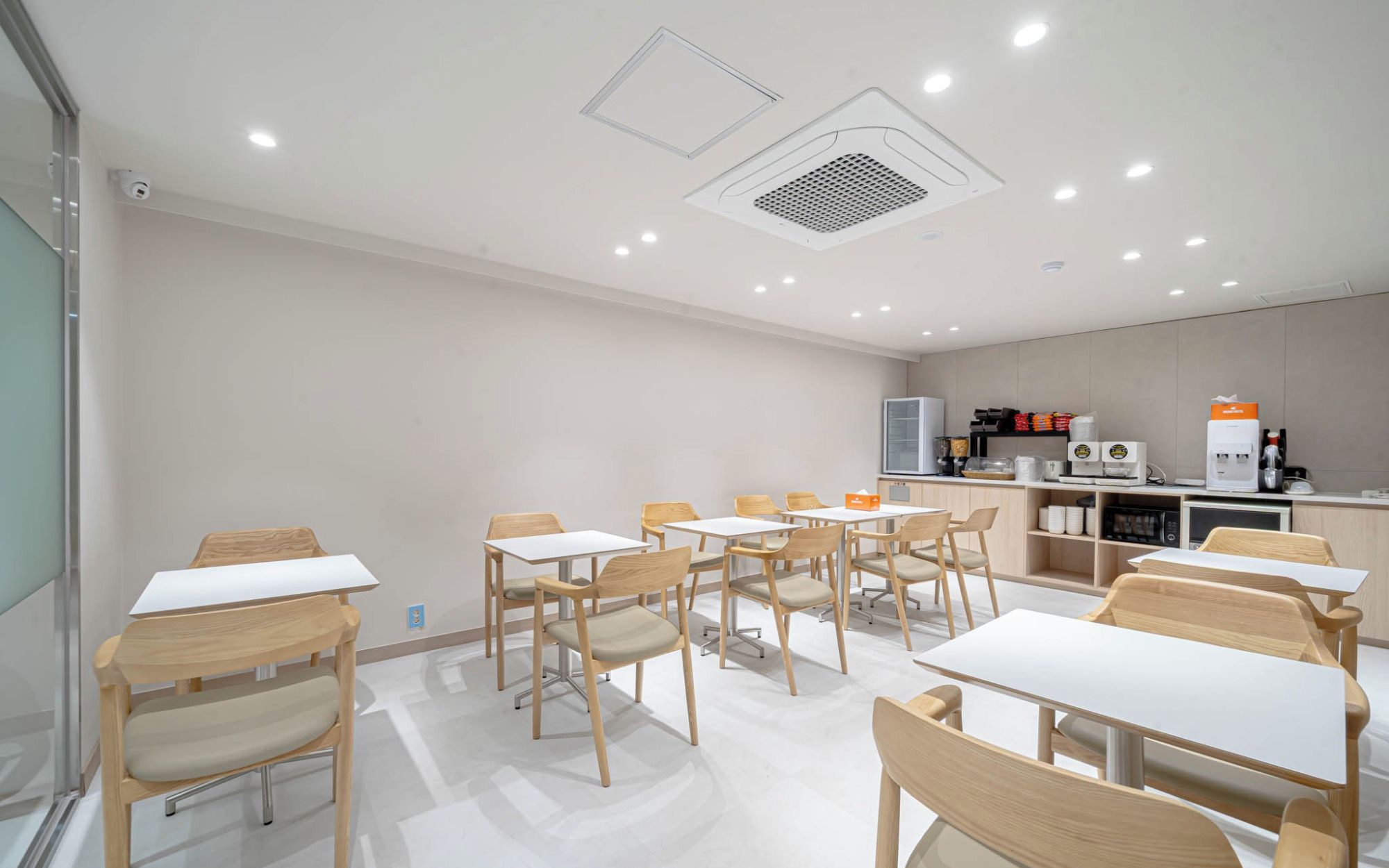 A modern dining area with multiple tables and chairs, a kitchen area with appliances, and a ceiling with recessed lighting.