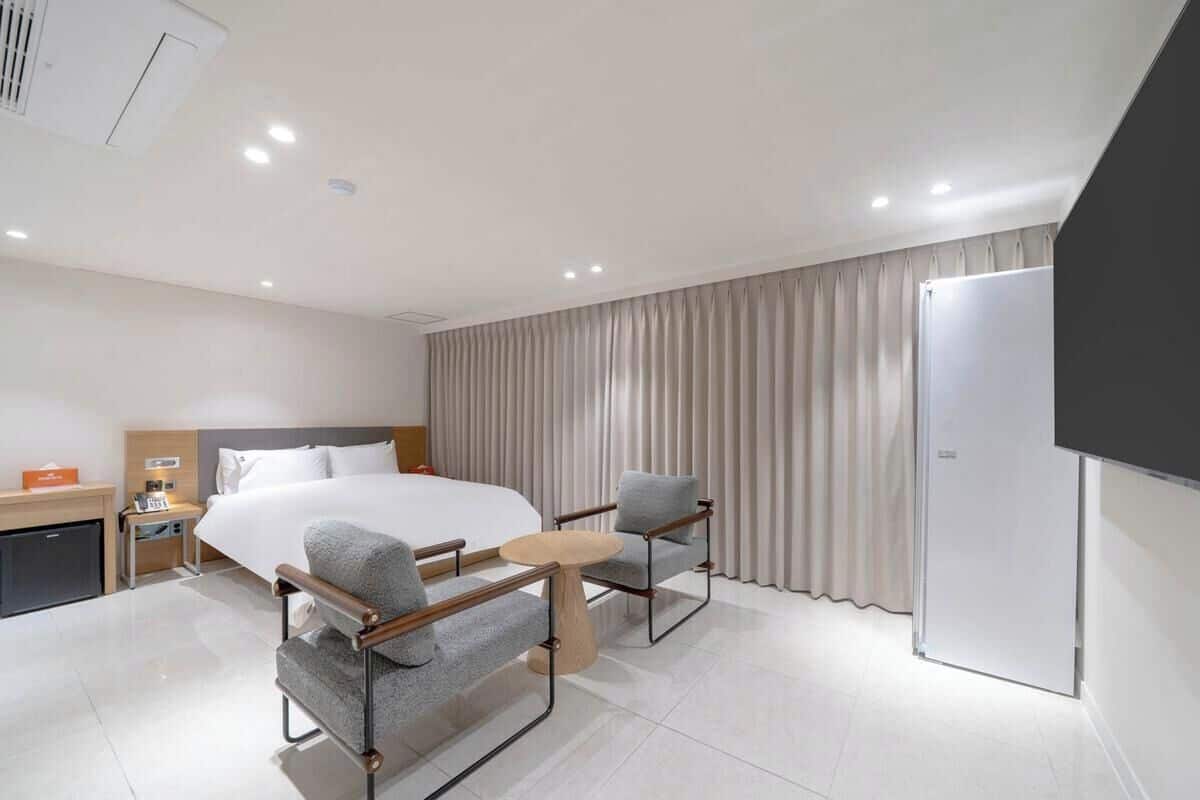 Una habitación de hotel moderna con una cama grande, dos sillones, una mesita redonda, un televisor y un armario.