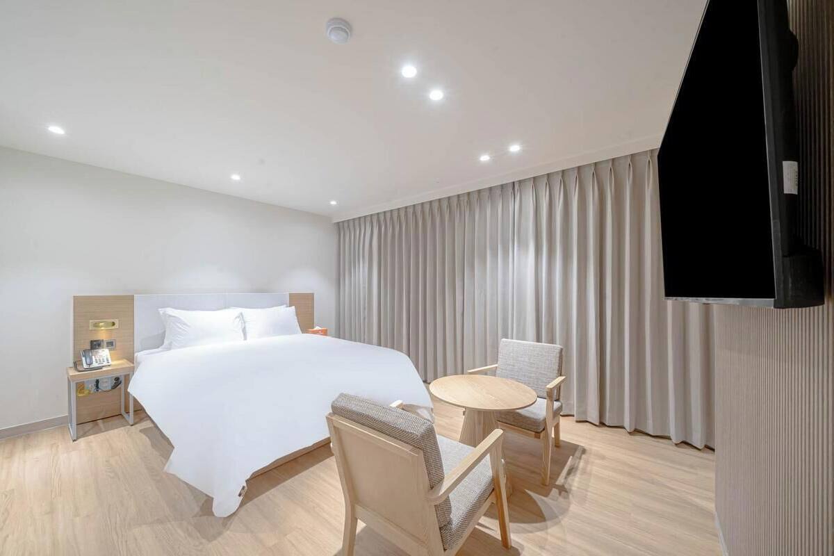 Una habitación de hotel con cama, mesita de noche, lámpara, silla, mesa redonda y cortinas.
