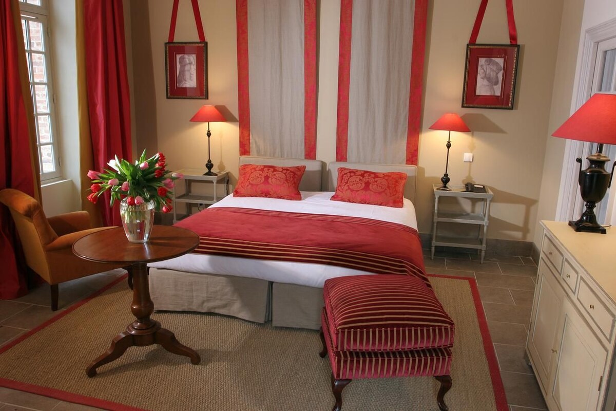 Habitación de hotel con cama, dos mesitas de noche, una silla, una mesita con un florero y un banco.