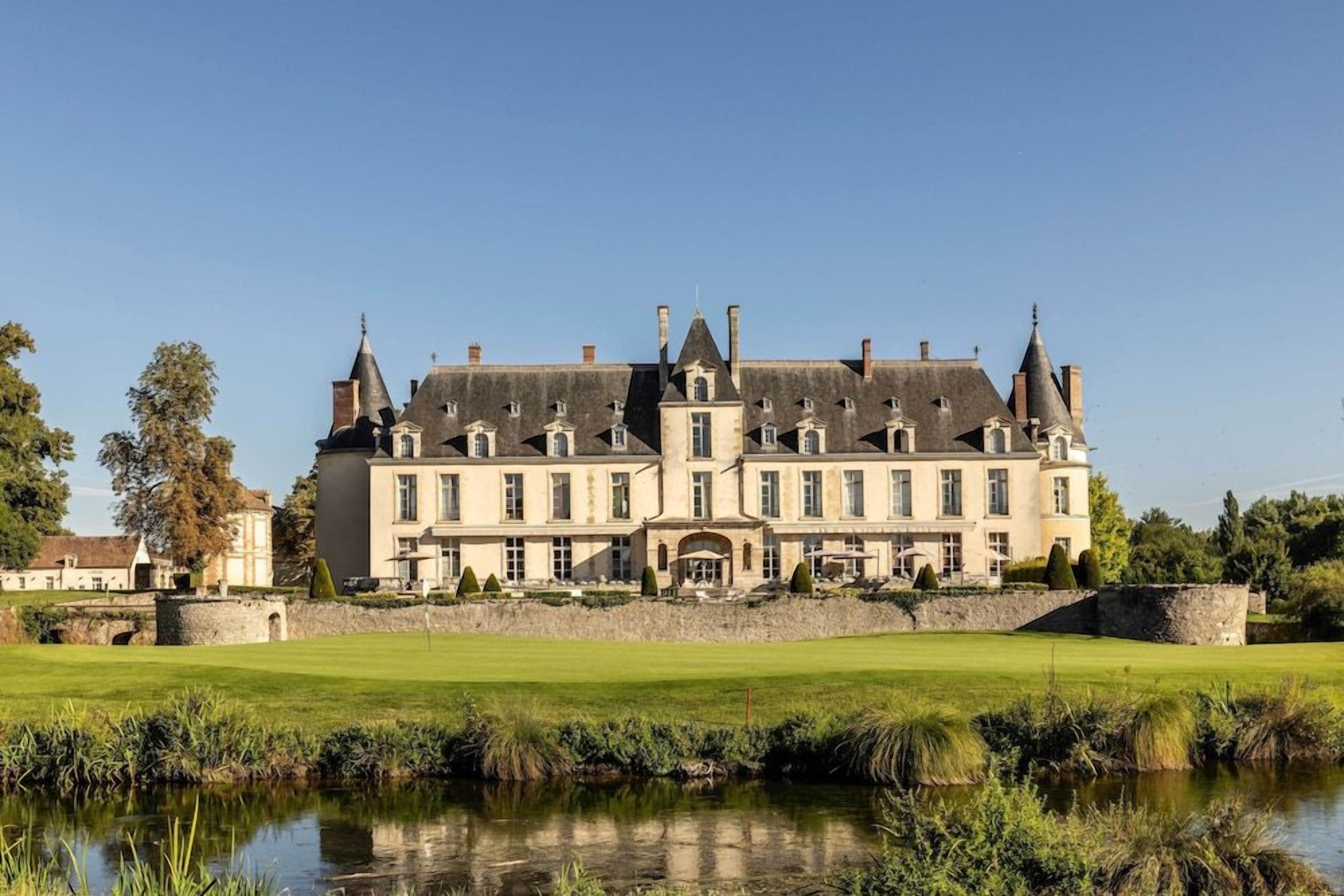 Un grand bâtiment historique, doté de nombreuses tours et d’une façade symétrique, entouré d’une pelouse bien entretenue et bordé d’un plan d’eau.