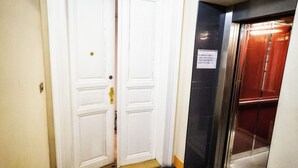 Een gang met een witte deur, een zwarte lift en een bord dat aangeeft dat de vuilnisbakken zich in de kelder bevinden.