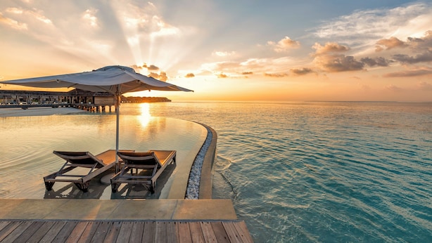 Ein Infinity-Pool mit Liegestühlen und Sonnenschirm, mit Blick auf ein ruhiges Meer bei Sonnenuntergang.