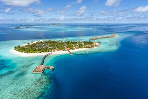 An aerial view of a tropical island resort with a white sandy beach, clear turquoise waters, and overwater bungalows connected by a bridge. - Hurawalhi Island Resort Maldives (Huruvalhi)