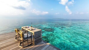 Eine Holzterrasse mit Tisch und Stühlen mit Blick auf klares türkisfarbenes Wasser und eine entfernte Insel.