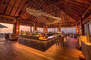 Een bar met een stenen toonbank, rieten meubels en uitzicht op de oceaan tijdens de zonsondergang.