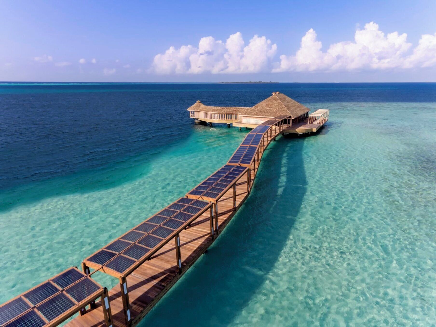 A wooden overwater walkway leading to a bungalow with a thatched roof, connected by a floating structure.