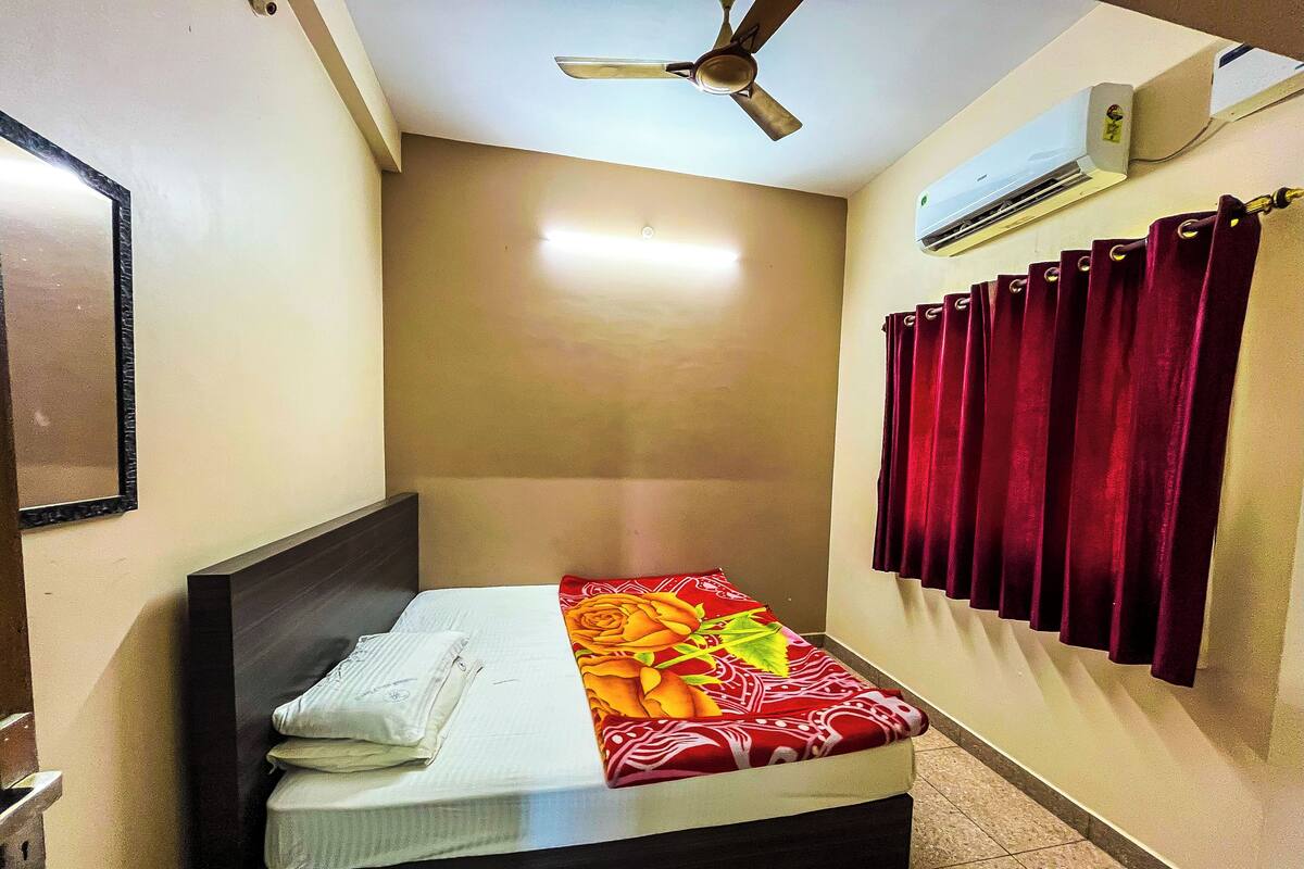 A small, single-bed room with a ceiling fan, an air conditioning unit, and a window with red curtains.
