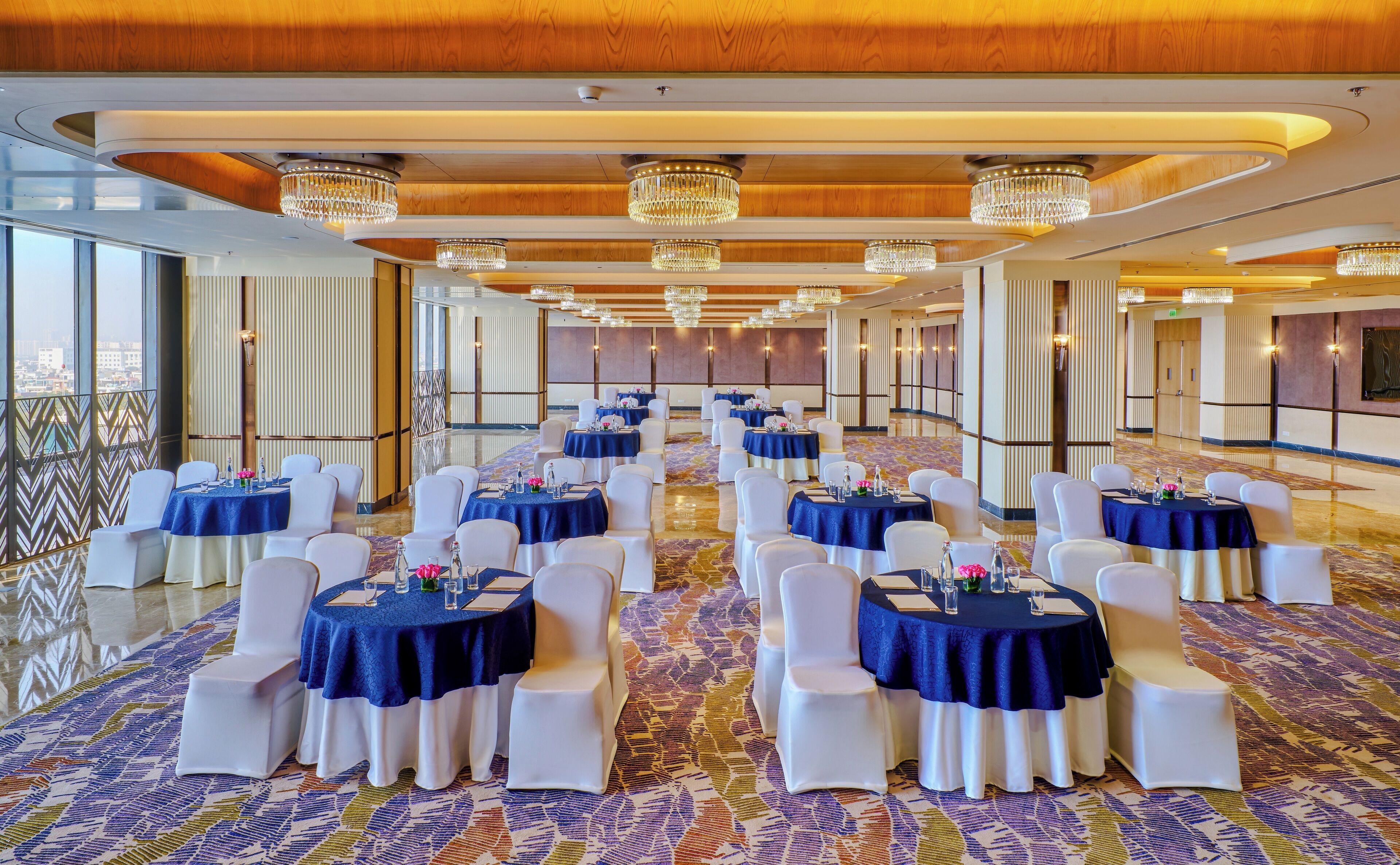 A spacious banquet hall with round tables, white chairs, and chandeliers.