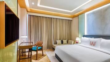 Quarto de hotel com cama, escrivaninha, cadeira, televisão e janela com cortinas.