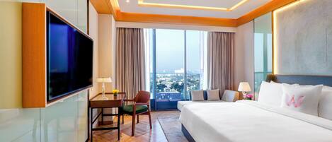 A hotel room with a large bed, a flat-screen TV, a desk with a chair, and a view of the city through large windows.