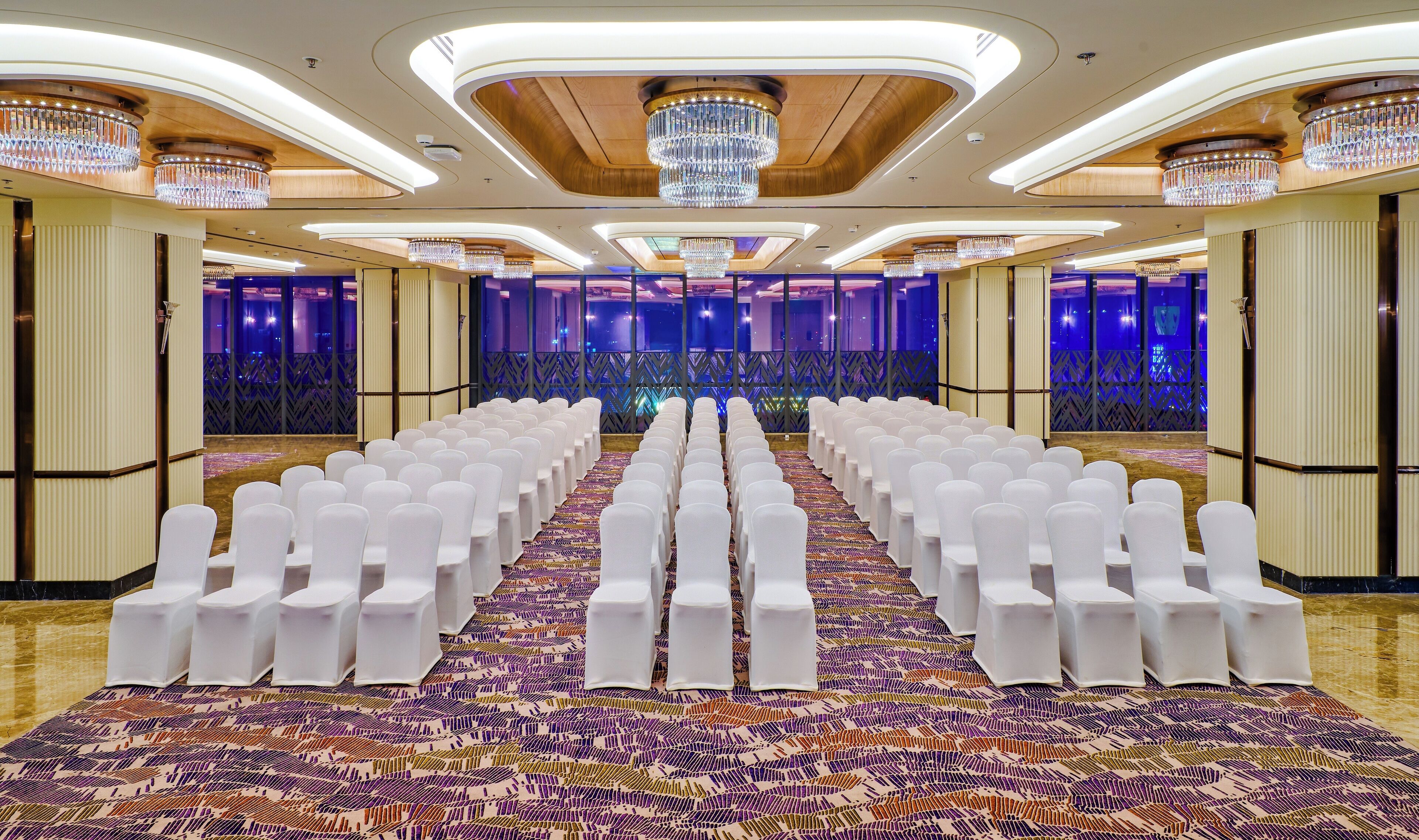 A conference room with rows of white chairs, a patterned carpet, and chandeliers.