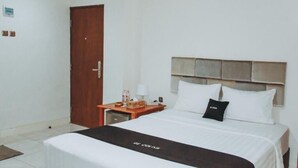 Habitación de hotel con cama, puerta de madera, mesita de noche con lámpara y una pequeña mesa con una caja de pañuelos y botellas.