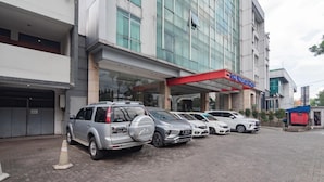 A hotel with a modern design, featuring a red awning and a glass facade. Several cars are parked in front, and a traffic cone is visible on the ground. - Urbanview Hotel Newton Riau Bandung (Bandung)