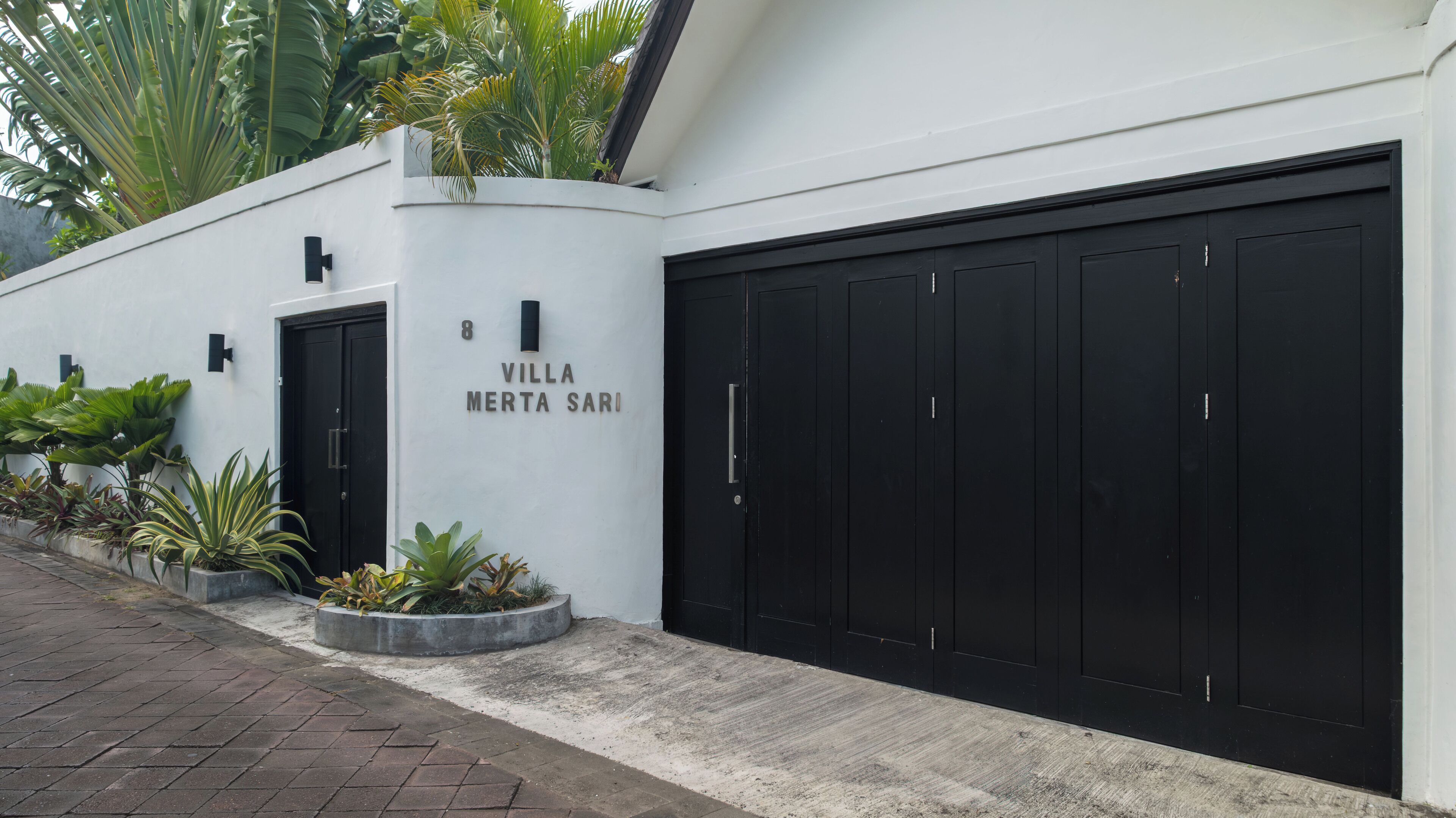 A modern villa with a black garage door, a white exterior, and a nameplate reading "Villa Merta Sari".