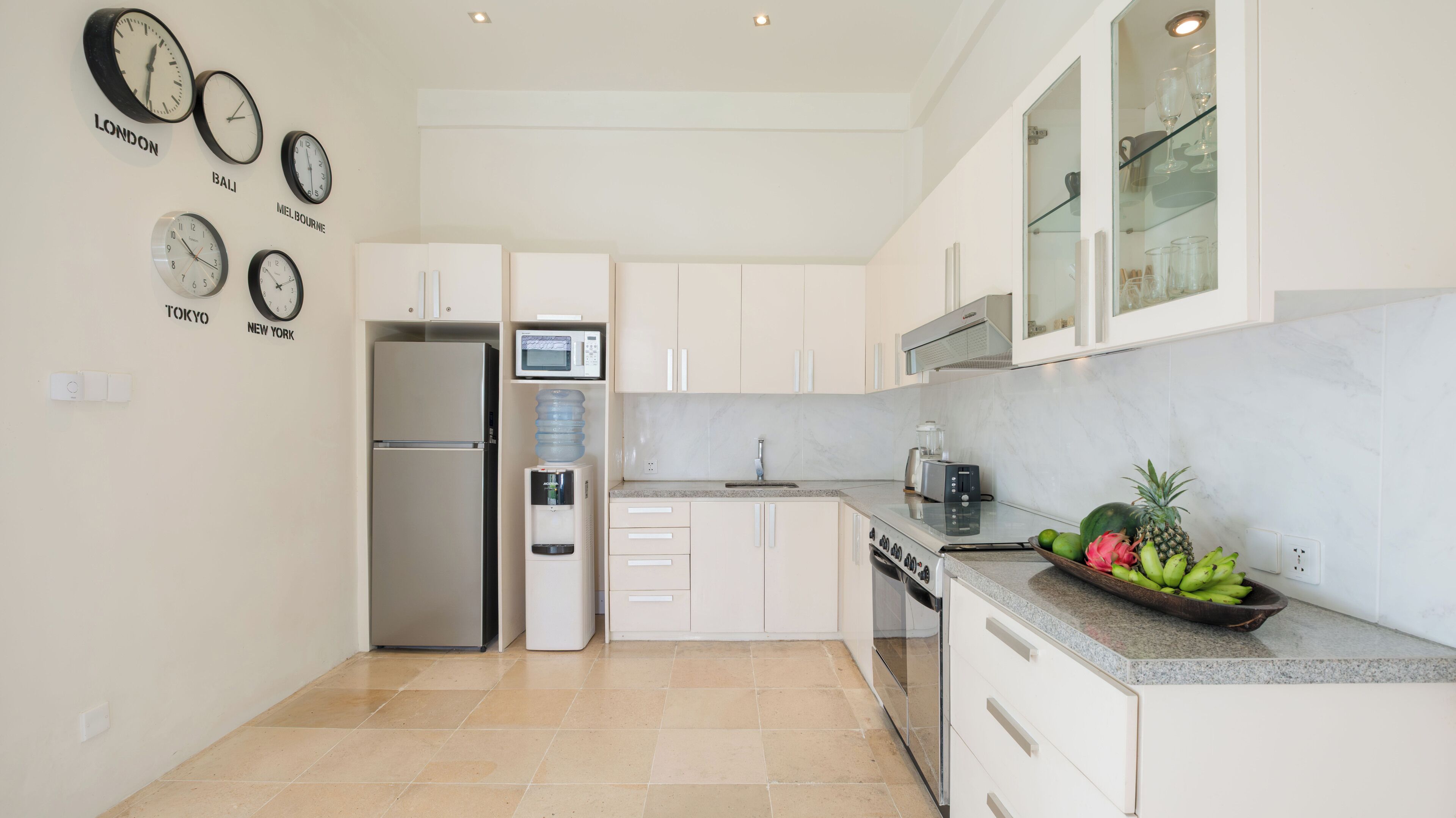 Una cucina moderna con mobili bianchi, frigorifero integrato, forno a microonde e dispenser d'acqua. Alle pareti, orologi che mostrano diversi fusi orari.