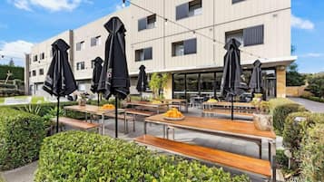 An outdoor seating area with tables and umbrellas, a building with multiple windows, and a well-maintained garden.