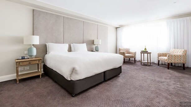 A hotel room with a large bed, two bedside tables, a lamp, and a chair. - Miramare Gardens Boutique Accommodation (Sydney)