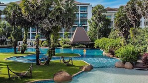 Zona de piscina de un resort con varias piscinas, palmeras y un edificio con balcones al fondo.