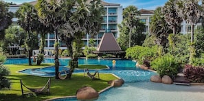 Zona de piscina de un resort con varias piscinas, palmeras y un edificio con balcones al fondo.