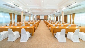 Un salón de conferencias con filas de mesas y sillas, piso alfombrado y ventanas amplias con cortinas.