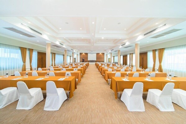 Une salle de conférence avec des rangées de tables et de chaises, un sol recouvert de moquette et de grandes fenêtres dotées de rideaux.