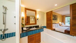 Un baño moderno con ducha, lavamanos y bañera. Hay un espejo grande sobre el lavamanos y se ve una cama a través de la puerta del baño.