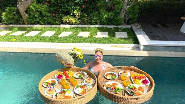 Montaje de comedor junto a la piscina con dos mesas de mimbre llenas de comida y bebidas, rodeado de exuberante follaje y una piscina cristalina.