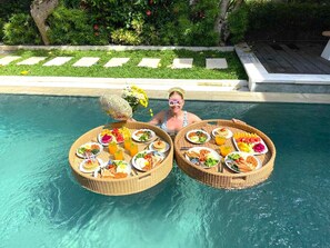 Ein Essbereich am Pool mit zwei Korbtischen, beladen mit Speisen und Getränken, umgeben von üppiger Bepflanzung und einem klaren Pool.