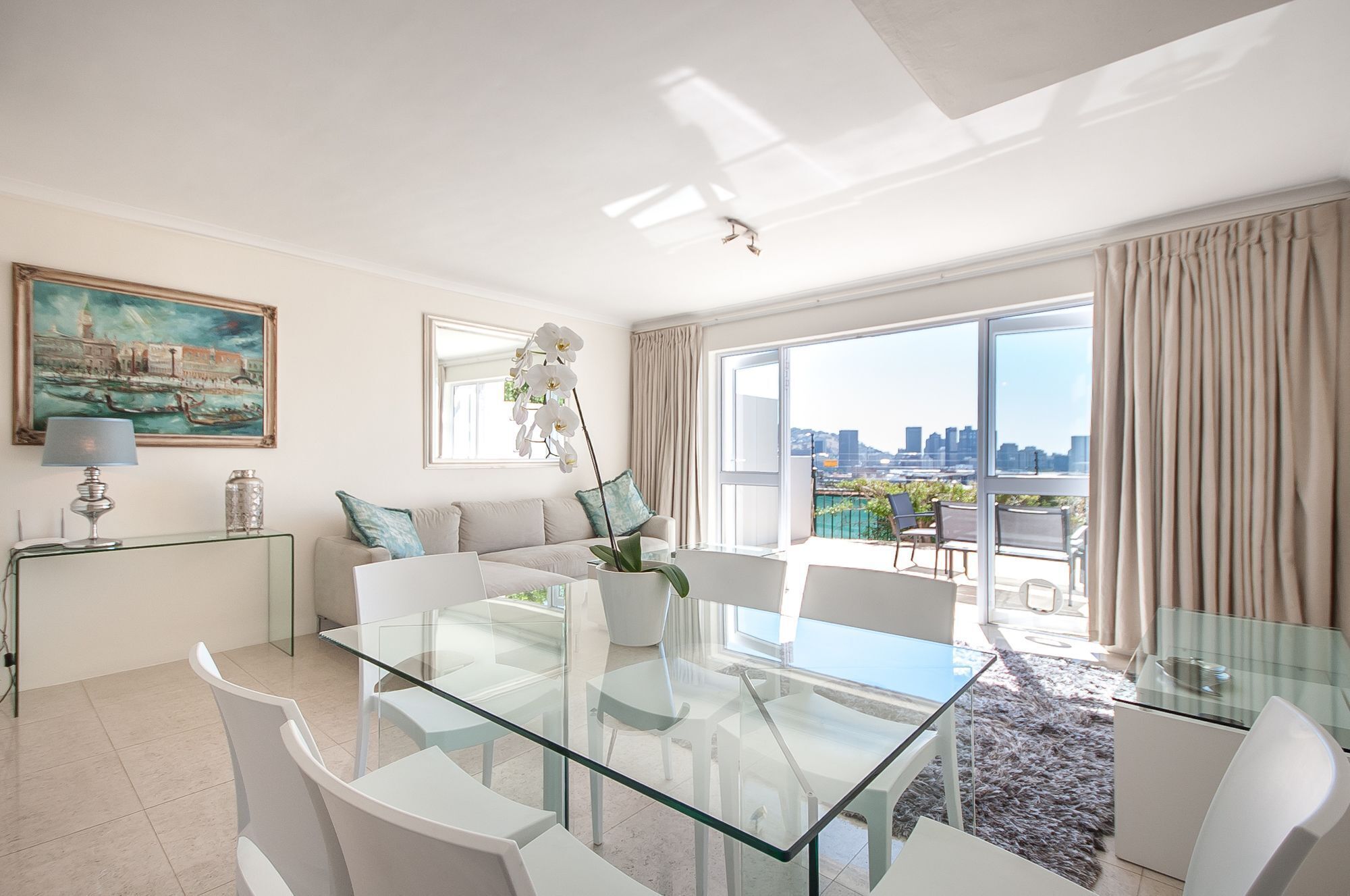 Ein modernes Wohnzimmer mit einem Glastisch, weißen Möbeln, Blick auf die Stadt und einem Gemälde an der Wand.