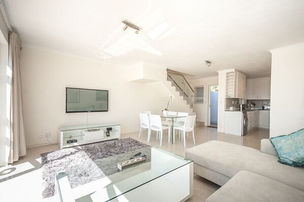 A modern living room with a glass coffee table, a sofa, a dining area with chairs, a kitchen with white cabinets, and a staircase. - 3 Canterbury Square (Cape Town)