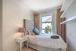 Une chambre avec une grande fenêtre, une tête de lit capitonnée, une table de chevet avec une lampe et un lit soigneusement fait.