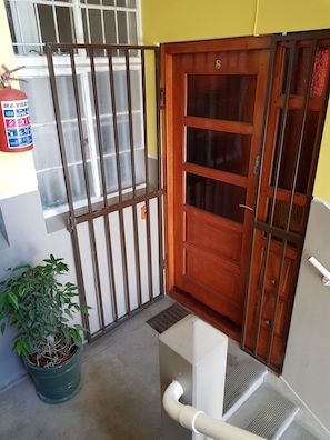 A room with a wooden door, a barred window, a potted plant, and a white pipe. - Bramber Court (Cape Town)