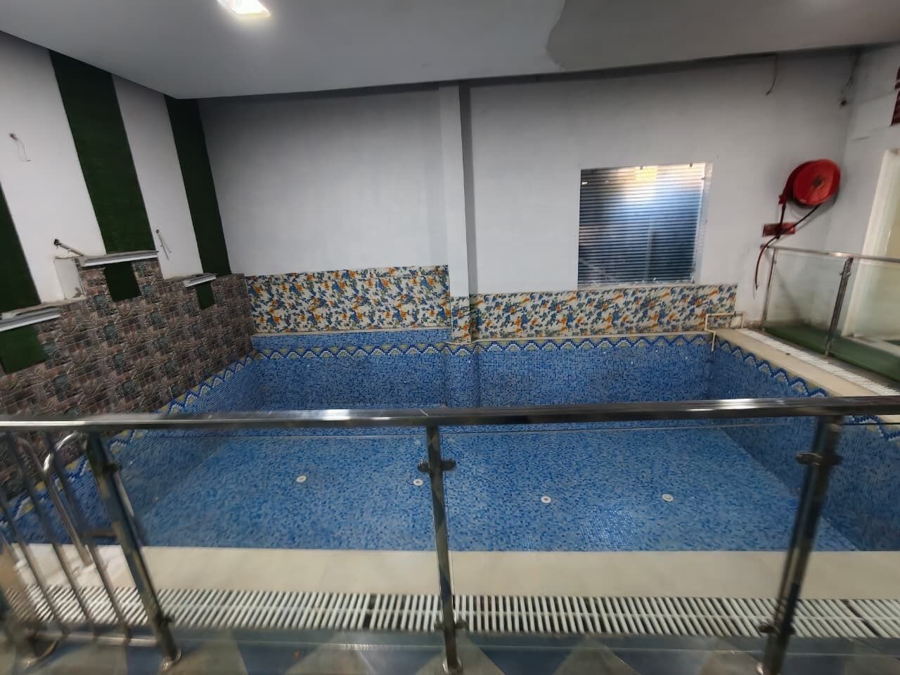 A swimming pool with a tiled border, a metal railing, and a window with blinds.