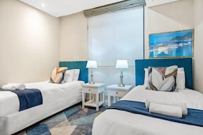 A hotel room with two beds, a window with a view, a painting on the wall, and a small table with lamps. - 236 Eden On the Bay (Cape Town)