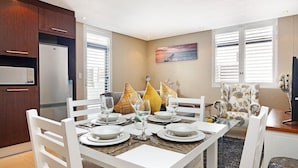 A dining area with a white table set for four, a refrigerator, and a window with blinds. - 236 Eden On the Bay (Cape Town)