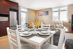 Un comedor con una mesa blanca para cuatro, un refrigerador y una ventana con persianas.