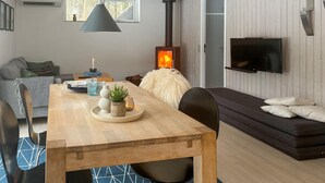 A cozy living room with a wooden dining table, a sofa, a TV mounted on the wall, and a wood-burning stove. - 2 bedroom beautiful home in øster assels (Øster Assels)