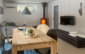 A cozy living room with a wooden dining table, a sofa, a TV mounted on the wall, and a wood-burning stove. - 2 bedroom beautiful home in øster assels (Øster Assels)