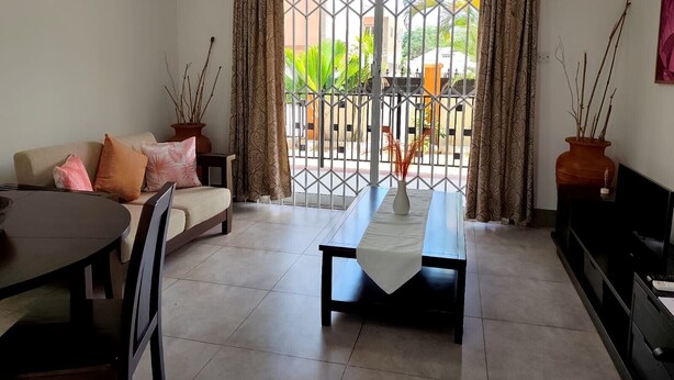 Una sala de estar con sofá, una mesa de comedor y televisor. Hay un balcón con una reja de metal y cortinas.